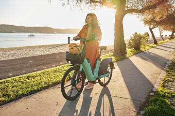 Pregnant woman in orange coat on electric bicycle on the promenade at sunset - obrazy, fototapety, plakaty