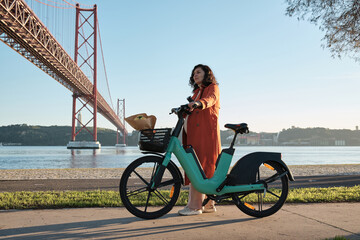 a Pregnant woman in orange coat on electric bicycle on the promenade in Lisbon near the bridge on...