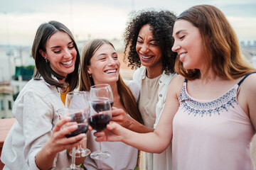 Group of happy young women friends having fun toasting wine glasses on a rooftop party, drinking and laughing together. Four cheerful girls smiling an clinking with drinks on a social reunion at bar