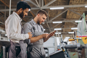 A male inspector or operator of a workshop for the production of aluminum and plastic wreaths trains an intern. International team of men working together near a machine in a factory.