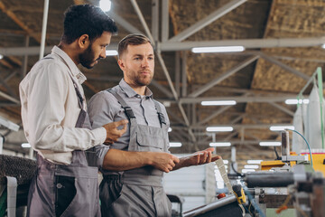 A male inspector or operator of a workshop for the production of aluminum and plastic wreaths trains an intern. International team of men working together near a machine in a factory.