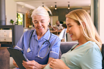 Senior Midwife With Digital Tablet Visiting Pregnant Woman At Home With Family In Background