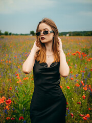 Beautiful young girl in a black evening dress and sunglasses posing against a poppy field on a cloudy summer day. Portrait of a female model outdoors. Rainy weather. Gray clouds. Vertical shot.