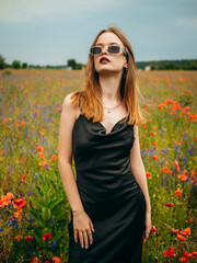 Beautiful young girl in a black evening dress and sunglasses posing against a poppy field on a cloudy summer day. Portrait of a female model outdoors. Rainy weather. Gray clouds. Vertical shot.