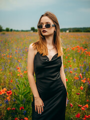 Beautiful young girl in a black evening dress and sunglasses posing against a poppy field on a cloudy summer day. Portrait of a female model outdoors. Rainy weather. Gray clouds. Vertical shot.