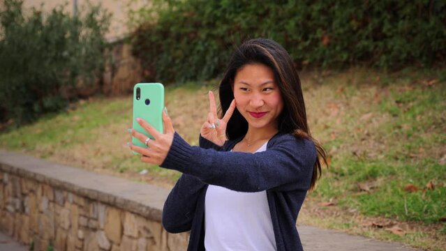 Cheerful asian student girl taking a selfie portrait with her smartphone device at university campus. Young woman having fun using a cellphone social media app. Teenage female holding a mobile phone