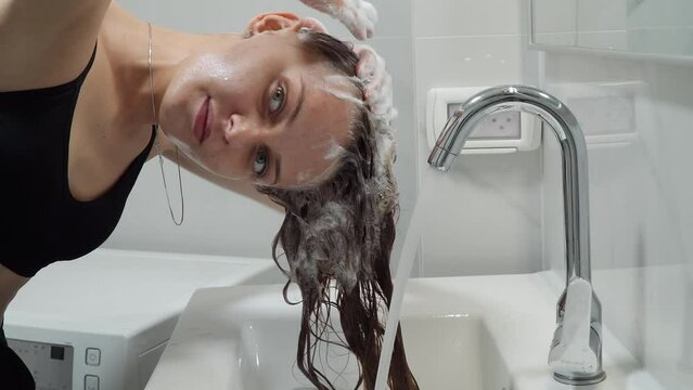 Caucasian girl washes her head with soap in the bathroom in the sink. European woman in a sports top bra washes her blond hair with shampoo with foam under a tap with water at home in an apartment