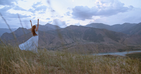 Woman traveler in light dress on mountain summit enjoying nature view with hands raised over clouds...