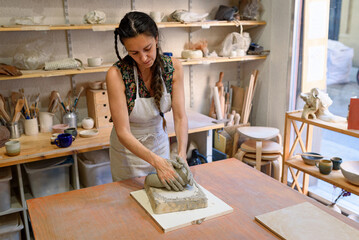 Young woman working raw ceramic and clay to make future ceramic product. Pottery workshop with natural light scraping, smoothing, shaping and sculpting. making clay pot