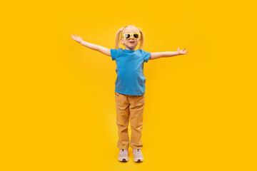 Smiling little girl in blue T-shirt and sunglasses raised her hands up. Portrait of child on yellow background.