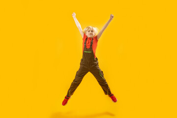 Fototapeta na wymiar Blond boy jumps high with his hands up. Portrait of schoolboy in overalls bouncing on yellow background. Copy space.