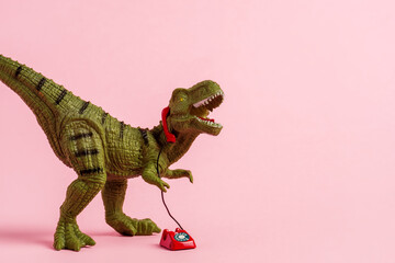Cute green toy dinosaur speaking on retro phone on pink background. Copy space.