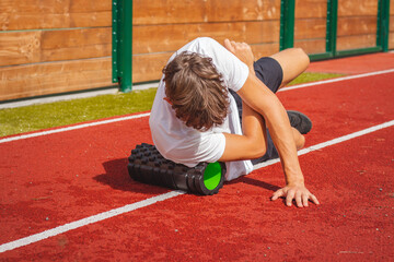 Brown-haired boy with an athletic physique on an athletic oval massages his muscles with a foam roller for better recovery. Post-workout