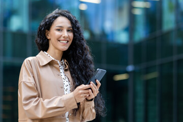 Portrait of a successful and satisfied Latin American business woman, female boss smiling and looking at the camera, holding a smartphone, using an online application, dialing a message and a call.
