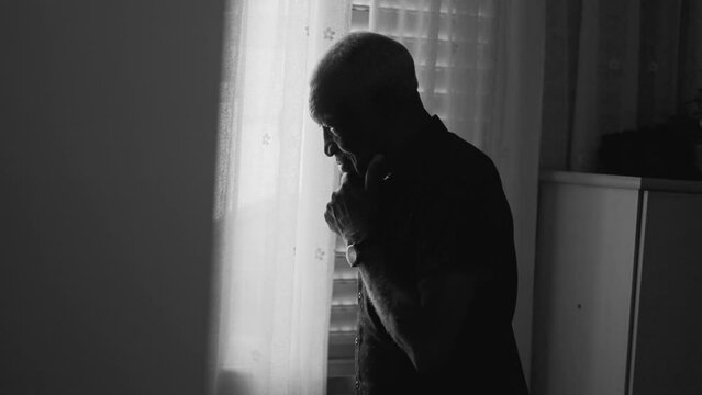 Dramatic scene of a black older man standing by window reflecting problems with hand in chin pondering solutions