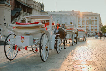 White fancy wooden horse drawn carriages on the Krakow main square or Rynek Glowny on a romantic sunny afternoon. Beautiful carriages ready to carry couples around. - obrazy, fototapety, plakaty