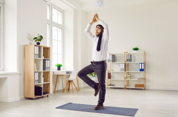 Funny young business man in white shirt, with necktie around his head practising stress management, taking break for relaxation, standing on yoga mat in office and doing vrikshasana aka tree pose