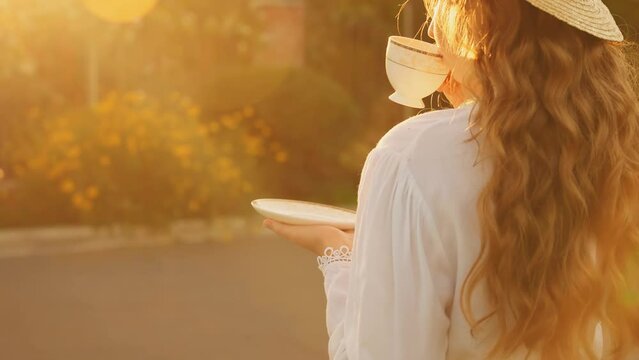 portrait Happy young vintage woman holding white cup in hands retro lady drinking tea back rear view. Fairy happy girl enjoy hot coffee drink. Green summer garden trees sunset magic sun light flare 4k