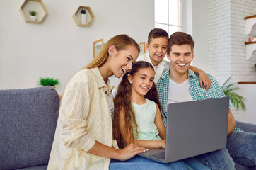 Parents together with their children enjoy weekend and laugh watching videos on computer at home. Happy young Caucasian family sitting on sofa in living room and looking at laptop screen.