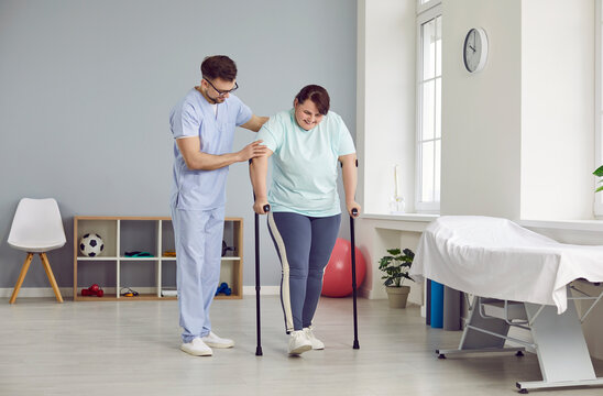 A young cheerful friendly nurse helping fat plus size female patient to walk with her crutches in medical clinic. Male physiotherapist or health care worker helping overweight woman in rehab.
