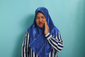 A middle-aged Asian woman in a blue hijab and striped shirt is suffering from a severe toothache while standing against a blue background