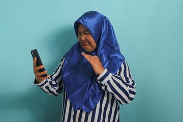 A middle-aged Asian woman, wearing a blue hijab and a striped shirt, is holding and looking at her...