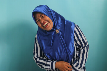 A funny Middle-Aged Asian woman, in a blue hijab and striped shirt, bursts into uncontrollable laughter, isolated on a blue background