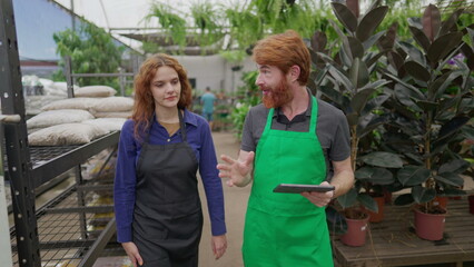 A male staff showing inventory to female colleague at local plant shop. Redhaired employees integrating technology with local business using tablet walking through isles