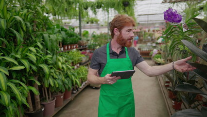 A male employee using tablet to browse inventory walking inside Flower Shop. Young man wearing green apron holding inspecting store with modern technology. Local business concept