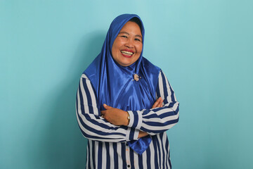 A smiling middle-aged Asian woman, wearing a blue hijab and a striped shirt, stands with her arms crossed, exuding happiness and confidence against a blue background