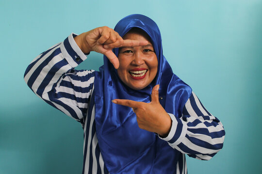 An inspired Middle-Aged Asian woman, in a blue hijab and striped shirt, makes a frame gesture, seeking the perfect angle or inspiration to capture a moment in a photo. Isolated on a blue background