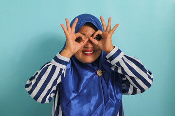 Middle-aged Asian woman in blue hijab, striped shirt, gesturing OK like binoculars, eyes peeking through fingers. Isolated on blue background.