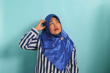 A confused middle-aged Asian woman in a blue hijab and striped shirt looks aside at the copy space, scratching her head while showing a doubting gesture. She is isolated on a blue background