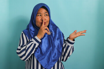A middle-aged Asian woman in a blue hijab points with her finger at the blank copy space aside. She makes a hush or be quiet gesture, telling a secret sale offer, isolated on blue background