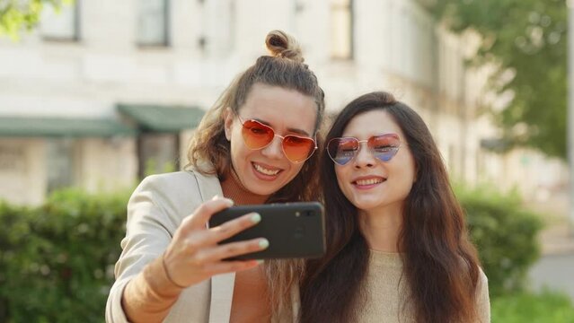 Two young smiling hipster blond women taking selfie self portrait photos on smartphone. Pretty stylish girls on street background. Positive emotions and love. LGBTQI, Pride Event, LGBT Pride Month
