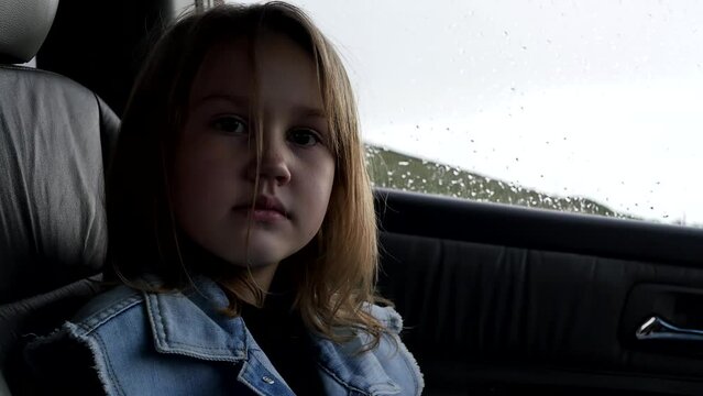 a cute European girl of seven years old dressed in a denim vest sitting inside the car looks. Child girl in a car with leather seats sitting in the passenger seat on a rainy day