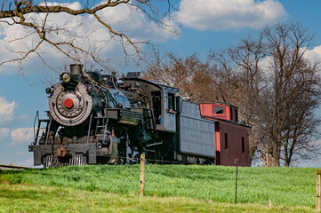 A View of a Restored Steam Passenger Train Traveling Thru Open Fields and Meadows on a Blue Sky with Clouds Day