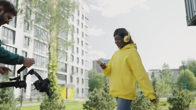 Shooting behind scenes, videographer shoots african-american girl in motion wearing headphones and yellow hoodie. Backstage. Slow motion