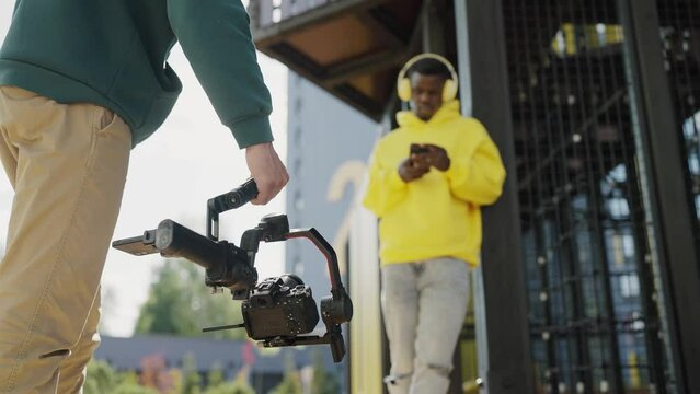 Professional videographer with camera on stabilizer shoots dark skinned young man with headphones and phone in his hands on territory of residential complex. Backstage. Slow motion