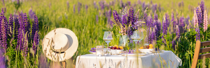 Beautiful countryside wedding of romantic dinner decor on blooming meadow with purple violet lupins flowers. Vase, wineglasses, cherry, apricot. Wooden table, basket. Sunset golden hour, summer banner