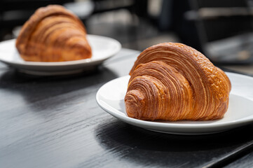 Two classic buttery croissants on white plates on a black wooden table at an outdoor cafe. French breakfast, bakery puff pastries against the backdrop of a summer veranda.