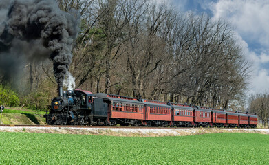 An Angled View of a Restored Steam Passenger Train Moving Slowly Blowing Lots of Black Smoke and White Steam on a Sunny Day