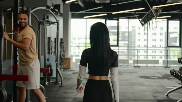 Back view sportswoman fitness trainer go through sport equipment in gym drink water after workout unknown woman meet man sportsman give high five respect two bodybuilders multiracial athletes greeting