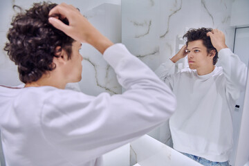 Young male looking at himself in bathroom mirror, touching curly hairstyle