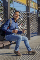 Young latin man sitting waiting for the bus with a paper cup of coffee in his hand.