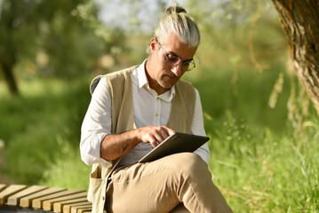 male business person drinking coffee talking on phone, doing analysis with tablet by the river in the park