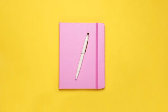 Closed pink notebook and pen on yellow background, top view