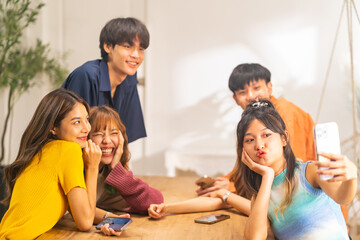 Group of Young Asian man and woman friends enjoy and fun reunion celebration party using mobile phone taking selfie together in living room at home on summer holiday vacation. Youth culture concept.