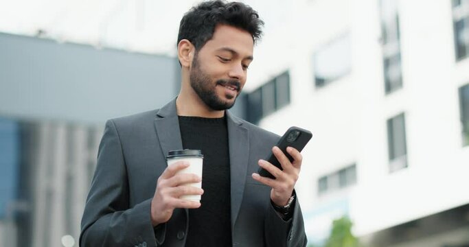 Good-looking corporate man walking while using a smartphone with cup of coffee. 
