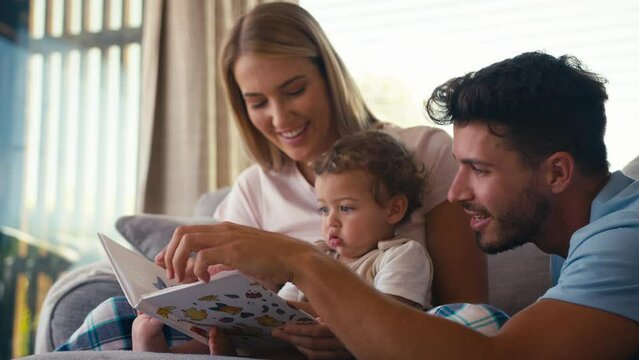 Family sitting on sofa at home with parents reading book with young son - shot in slow motion